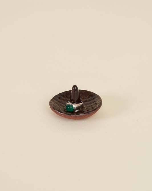 A silver ring with a large green stone sits around a pointed stem in the middle of a small ceramic ring holder dish with a checkerboard texture, made of a red stoneware and black amber glaze, sits on a cream background.