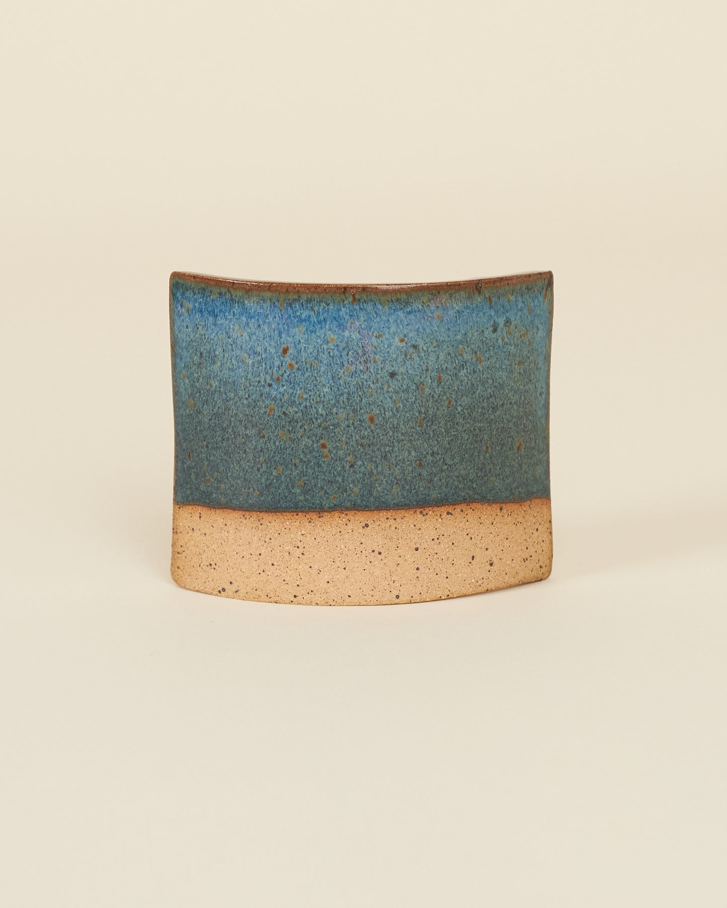A curved square test tile made of speckled tan clay and a rich glossy blue variegated glaze sits on a cream background.