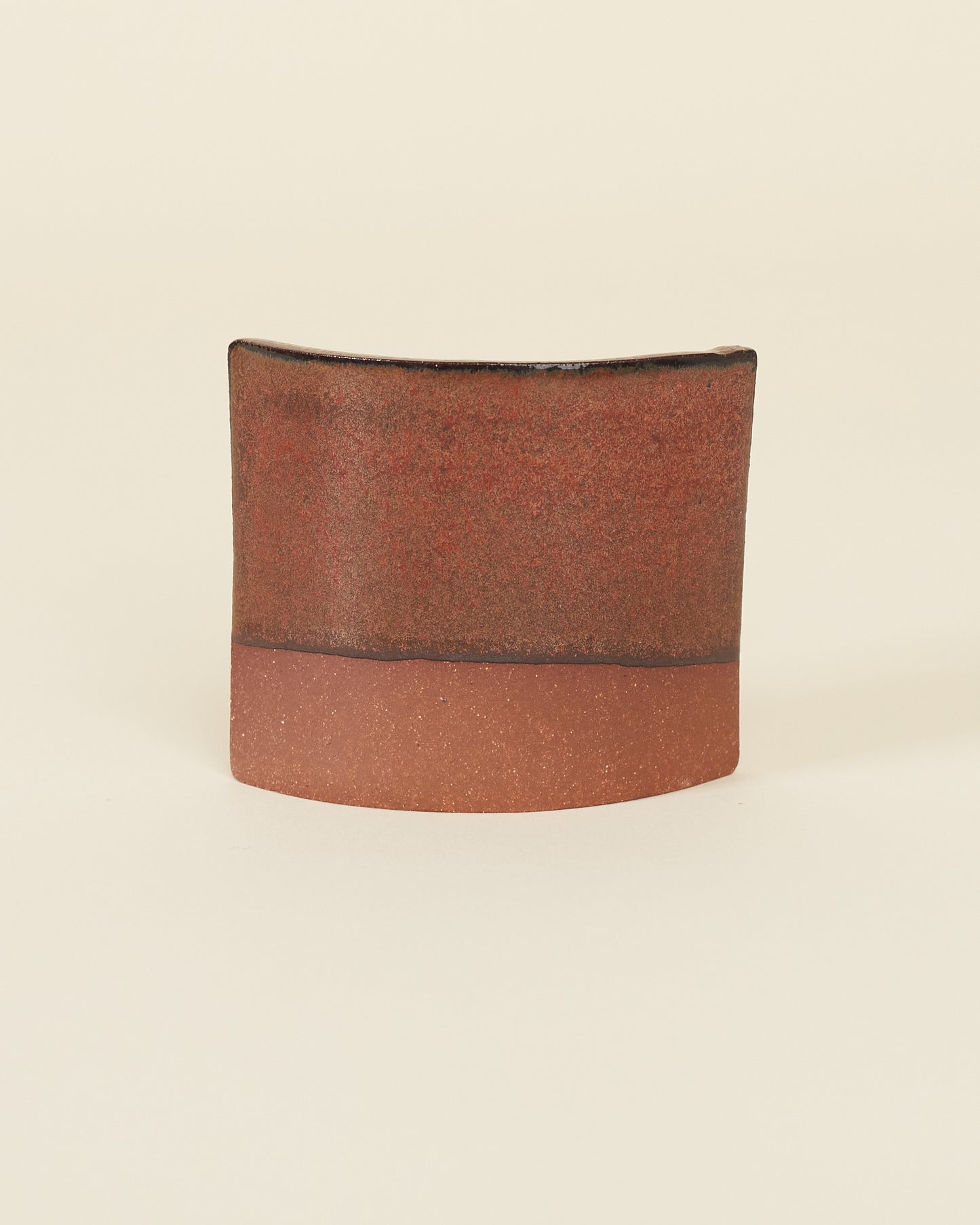 A curved square test tile made of red clay and a satin variegated red glaze that breaks black on the edges sits on a cream background.