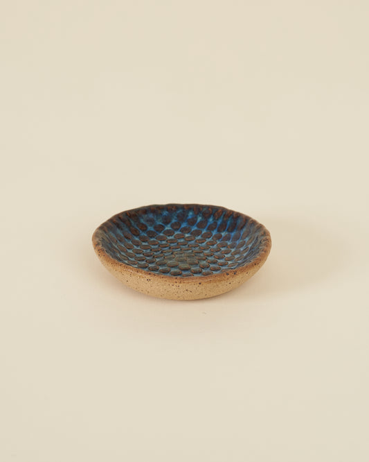 A small checkerboard textured dish made of speckled tan clay and a rich glossy blue variegated glaze sits on a cream background.