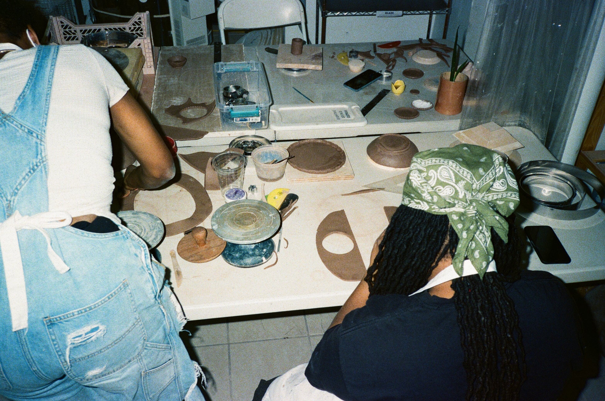 Grainy film image of two young women with dark brown skin and dreads from behind, one seated, one standing, working with slabs of clay at a table.