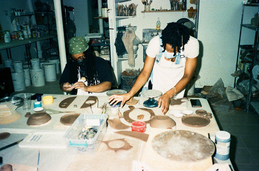 Grainy film image of two young women with dark brown skin and dreads, one seated, one standing, working with slabs of clay at a table.