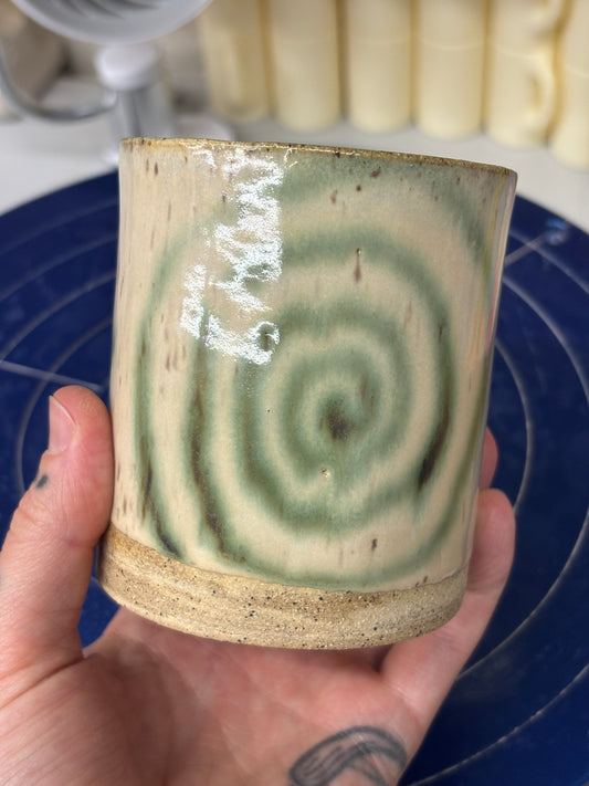Speckled ceramic cup with a cream glaze. Melty swirls and stars in green copper adorn the outside. Stamped with LKBK maker’s mark on the bottom.