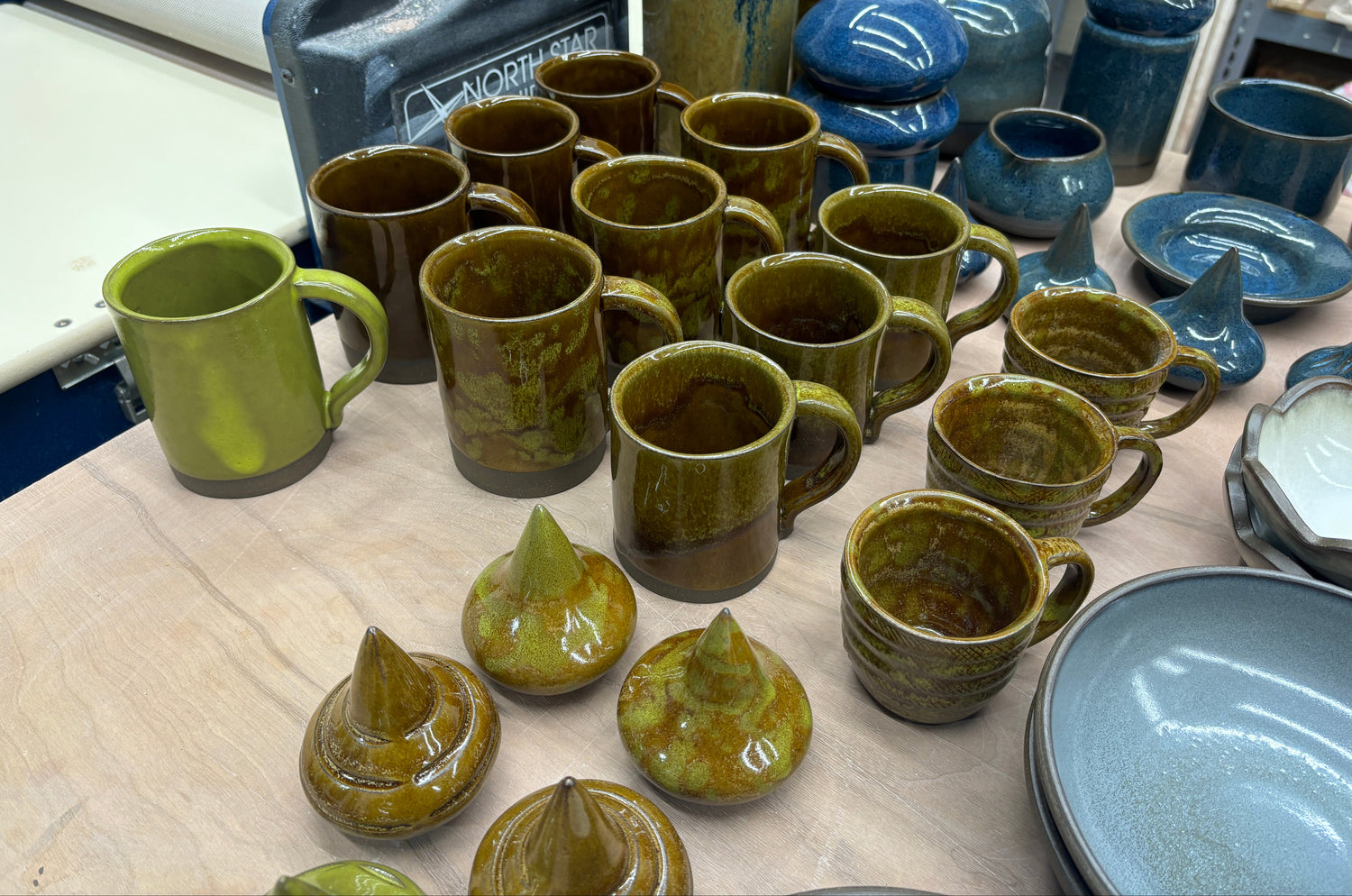 Collection of mugs, bowls, and vases in blues and greens on a wood table next to a slab roller in a ceramic studio.