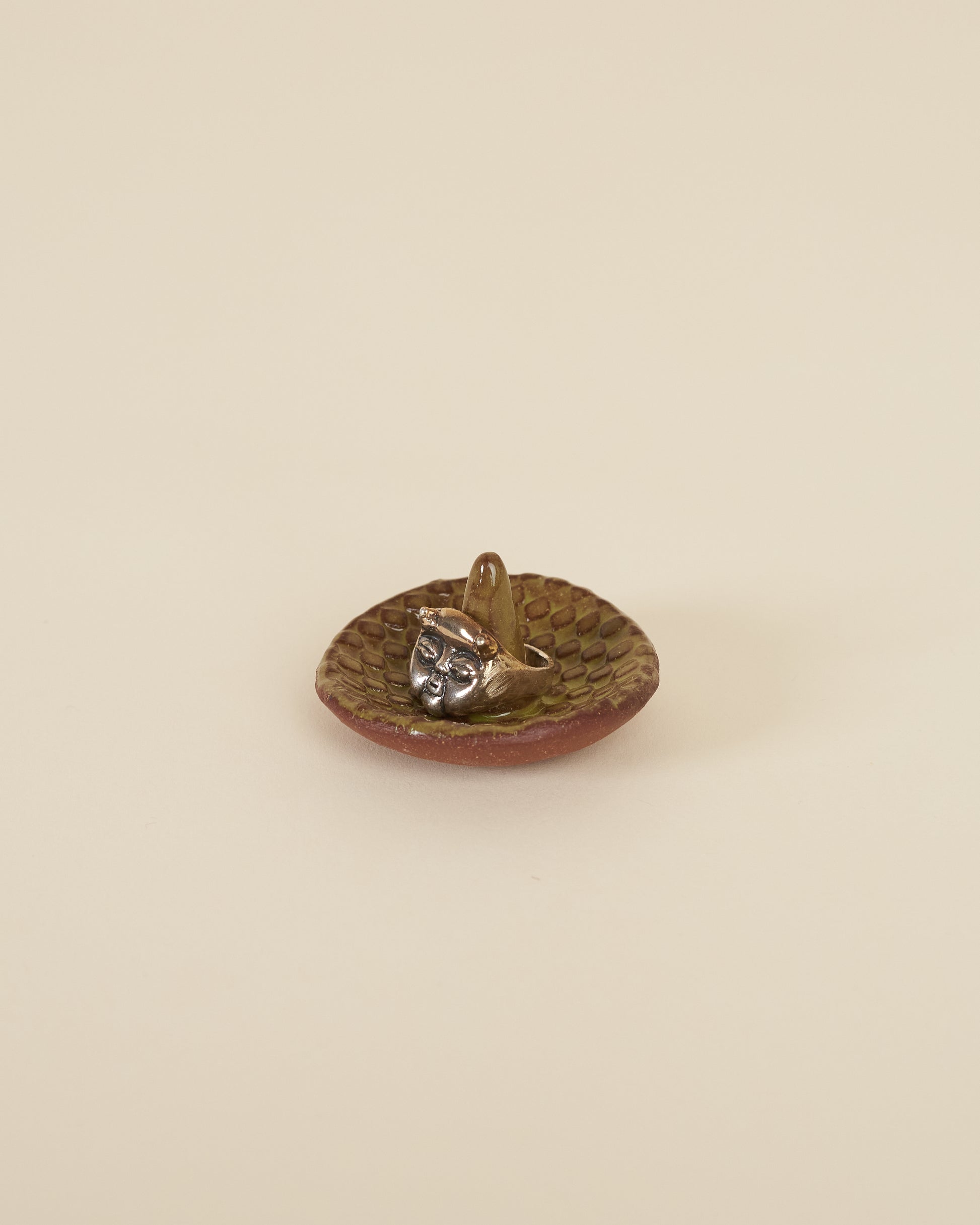 A large silver ring featuring a cherub face and small horns sits around a pointed stem in the middle of a small ceramic ring holder dish with a checkerboard texture, made of a red stoneware and black chartreuse glaze, sits on a cream background.