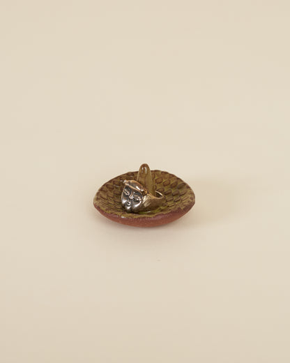 A large silver ring featuring a cherub face and small horns sits around a pointed stem in the middle of a small ceramic ring holder dish with a checkerboard texture, made of a red stoneware and black chartreuse glaze, sits on a cream background.
