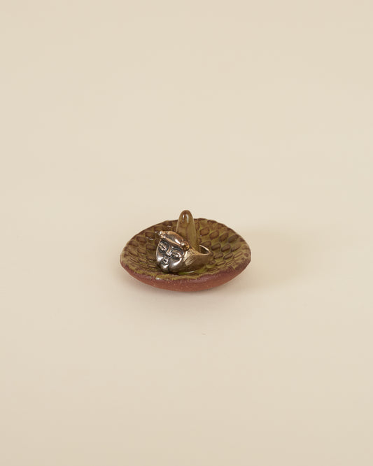 A large silver ring featuring a cherub face and small horns sits around a pointed stem in the middle of a small ceramic ring holder dish with a checkerboard texture, made of a red stoneware and black chartreuse glaze, sits on a cream background.