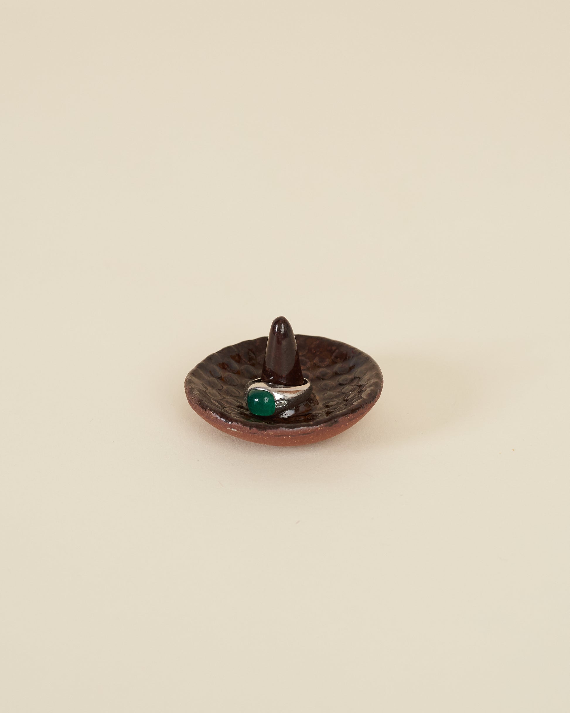 A silver ring with a large green stone sits around a pointed stem in the middle of a small ceramic ring holder dish with a checkerboard texture, made of a red stoneware and black amber glaze, sits on a cream background.