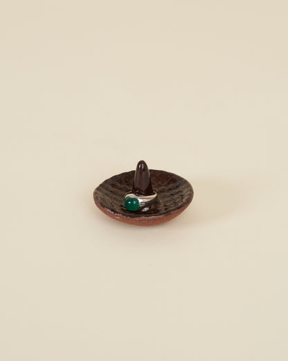 A silver ring with a large green stone sits around a pointed stem in the middle of a small ceramic ring holder dish with a checkerboard texture, made of a red stoneware and black amber glaze, sits on a cream background.