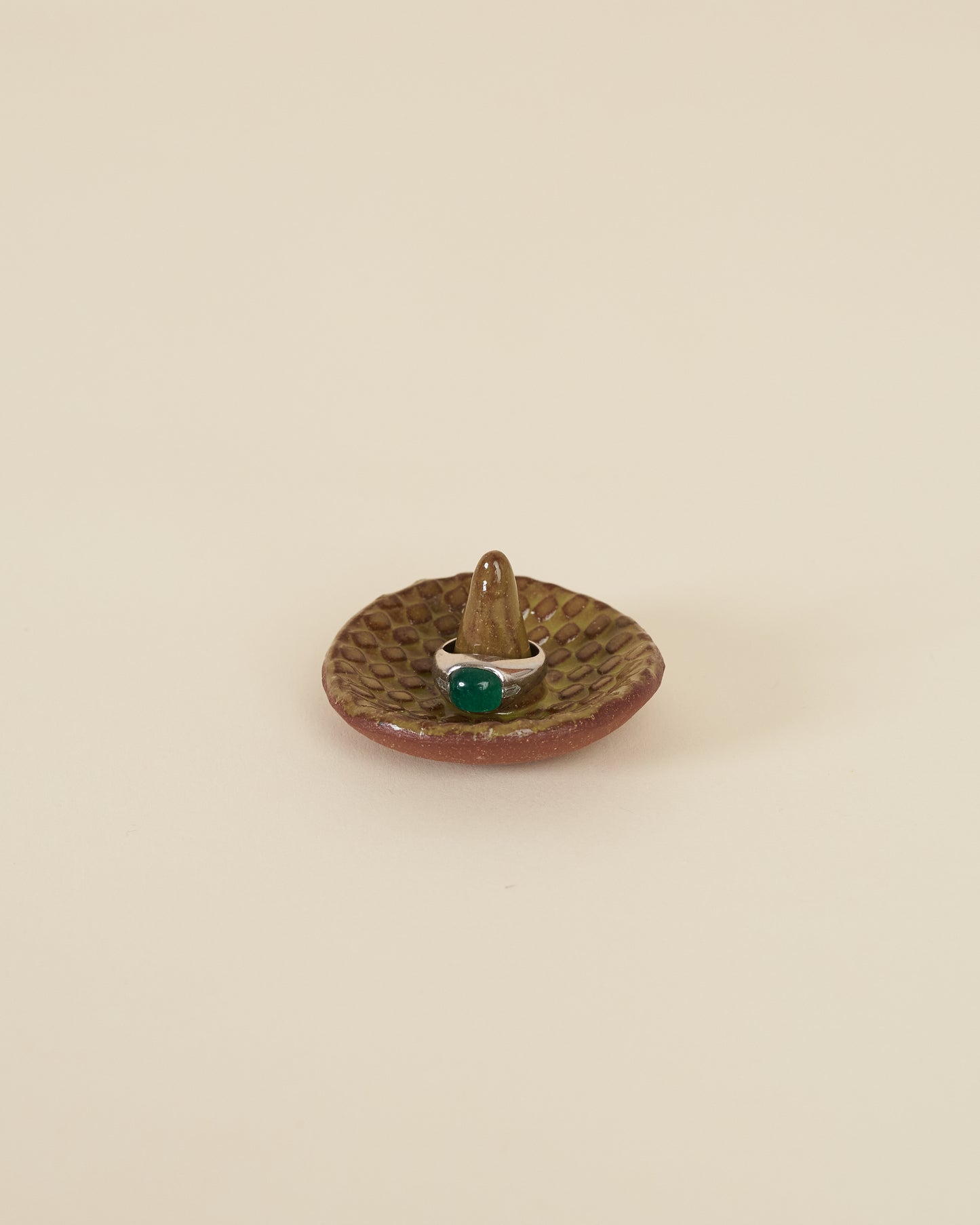 A silver ring with a large green stone sits around a pointed stem in the middle of a small ceramic ring holder dish with a checkerboard texture, made of a red stoneware and black chartreuse glaze, sits on a cream background.