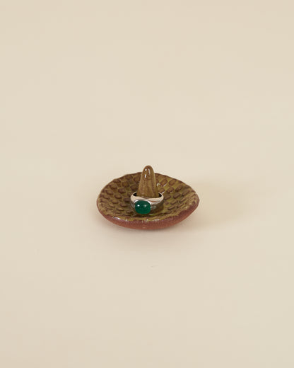 A silver ring with a large green stone sits around a pointed stem in the middle of a small ceramic ring holder dish with a checkerboard texture, made of a red stoneware and black chartreuse glaze, sits on a cream background.