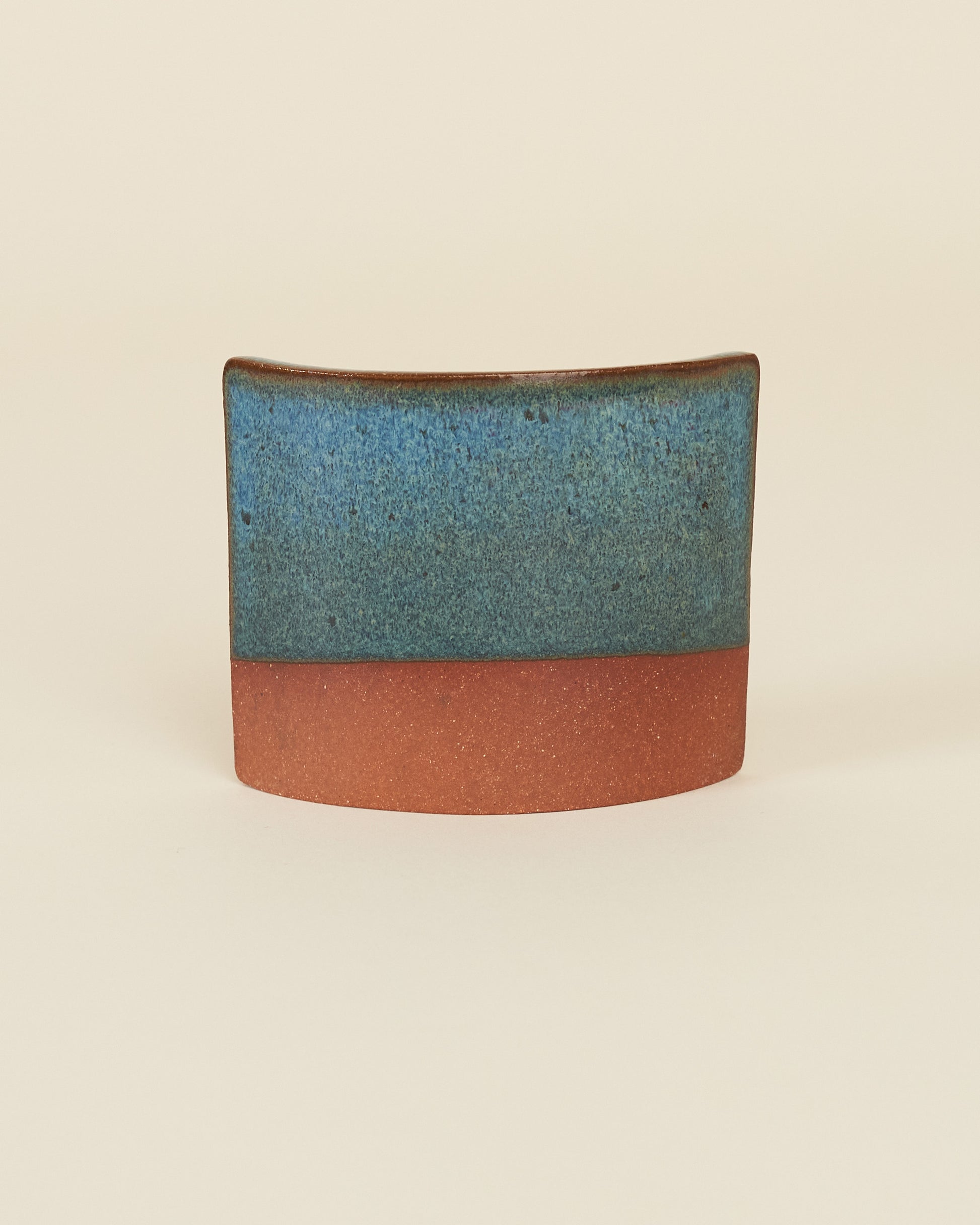 A curved square test tile made of red clay and a glossy rich blue variegated glaze sits on a cream background.