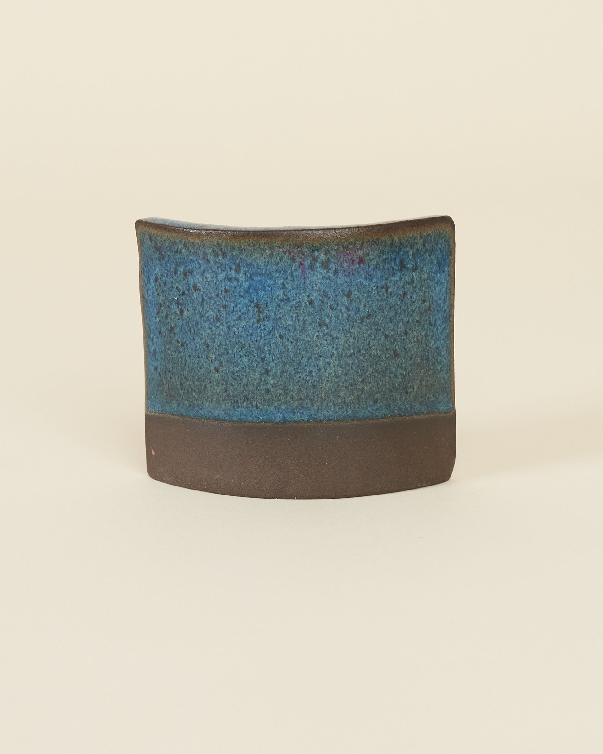 A curved square test tile made of dark brown clay and a rich glossy blue variegated glaze sits on a cream background.