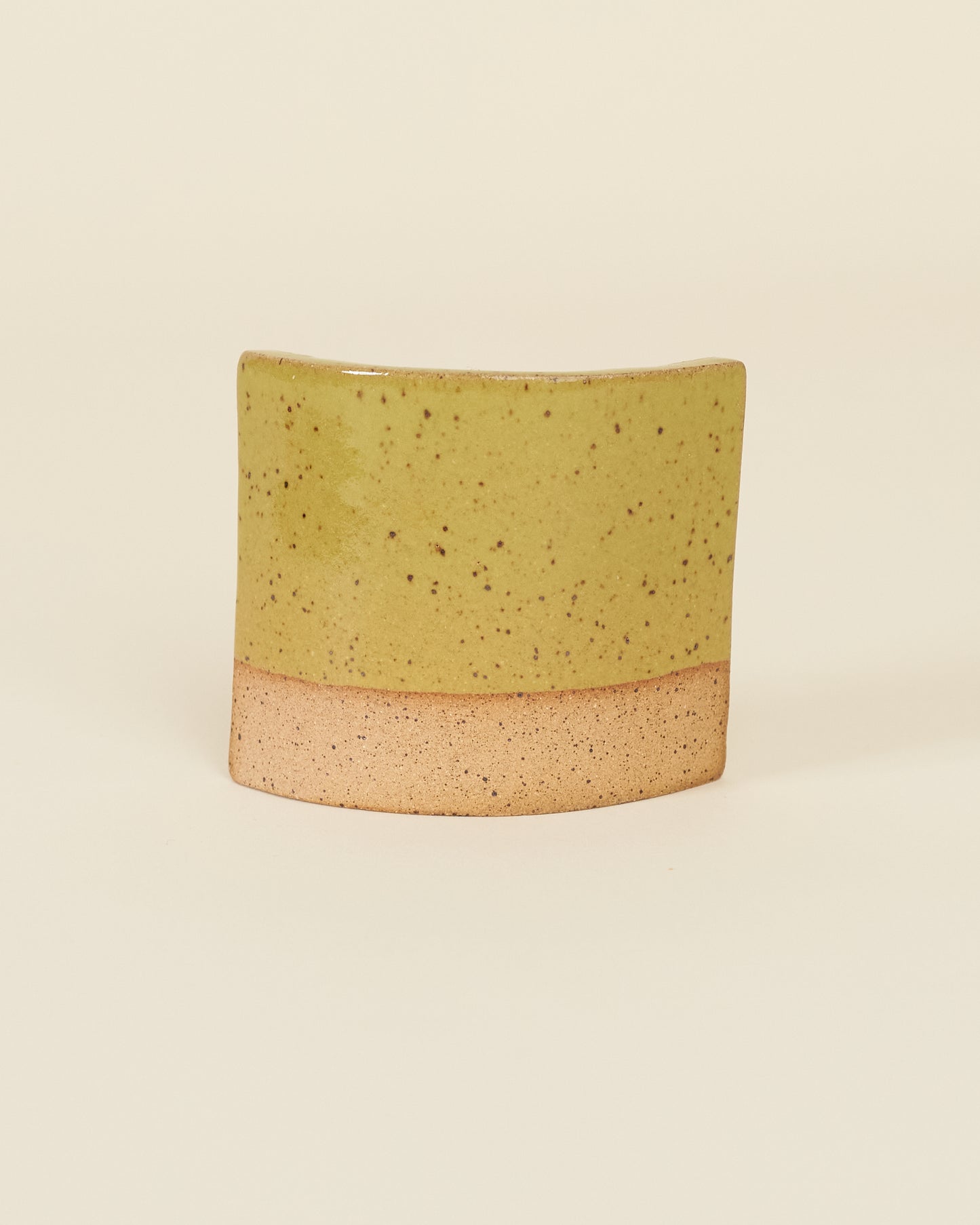 A curved square test tile made of speckled tan clay and a glossy chartreuse glaze sits on a cream background.
