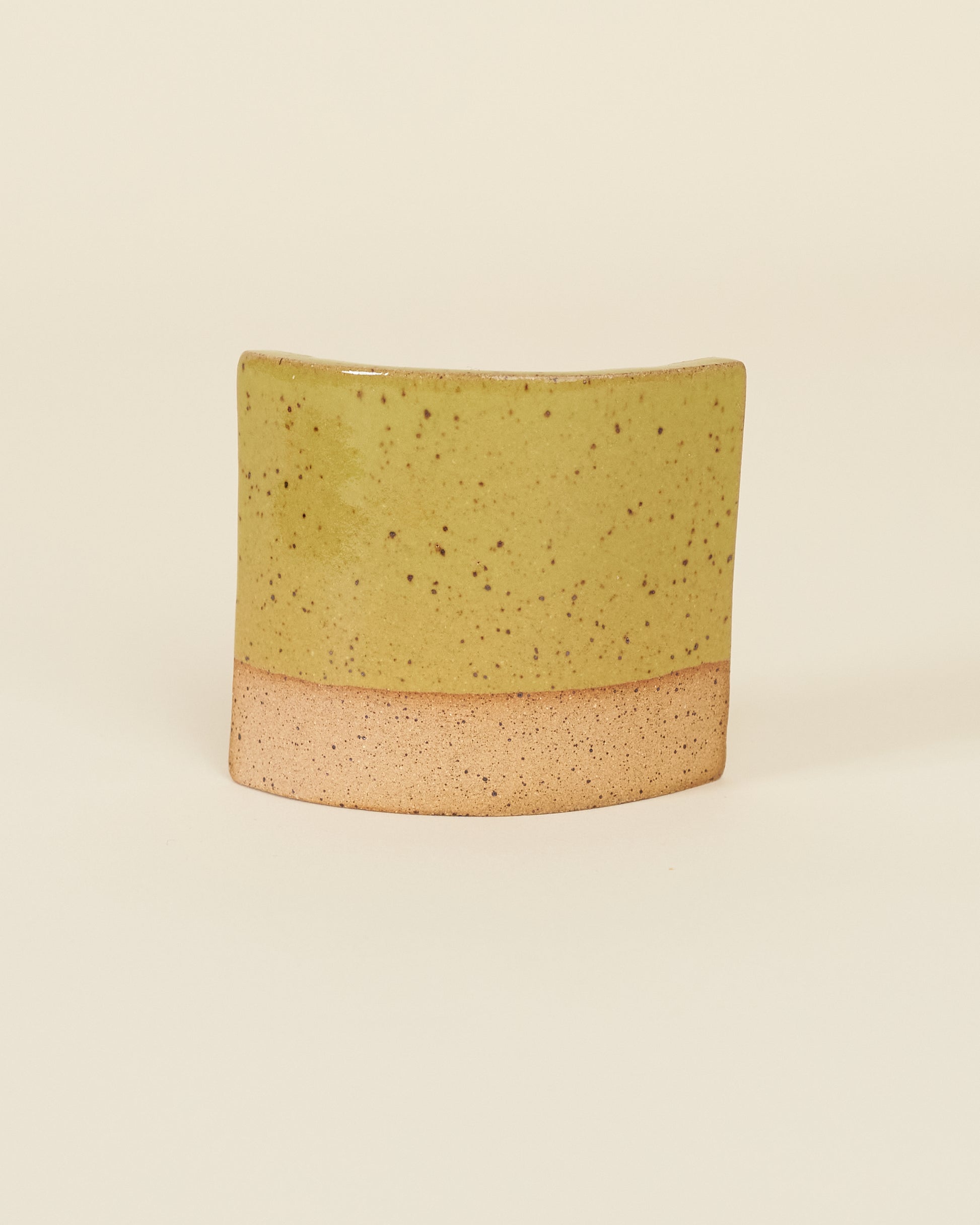 A curved square test tile made of speckled tan clay and a glossy chartreuse glaze sits on a cream background.
