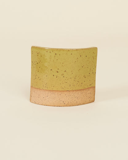 A curved square test tile made of speckled tan clay and a glossy chartreuse glaze sits on a cream background.