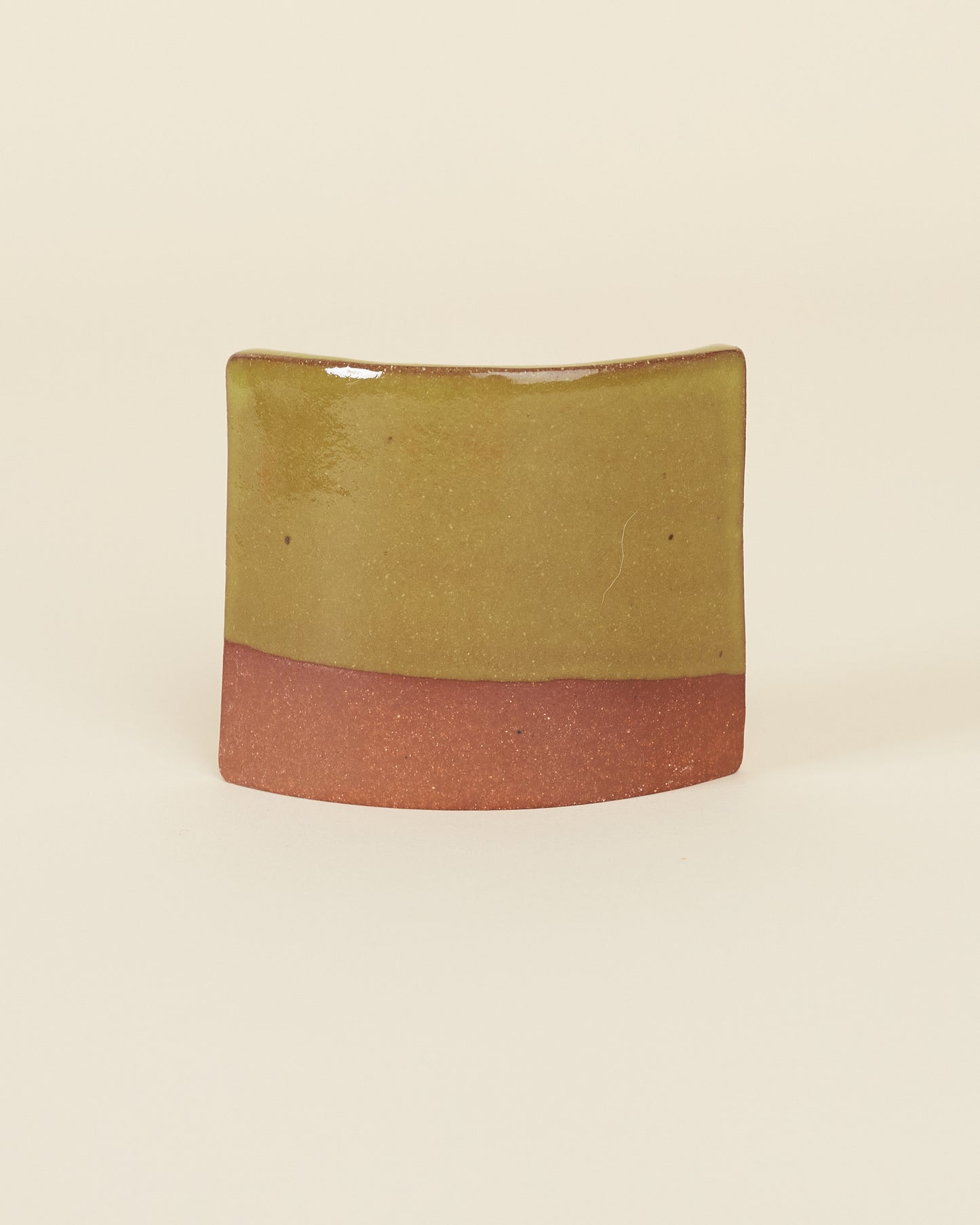 A curved square test tile made of red clay and a glossy chartreuse glaze sits on a cream background.