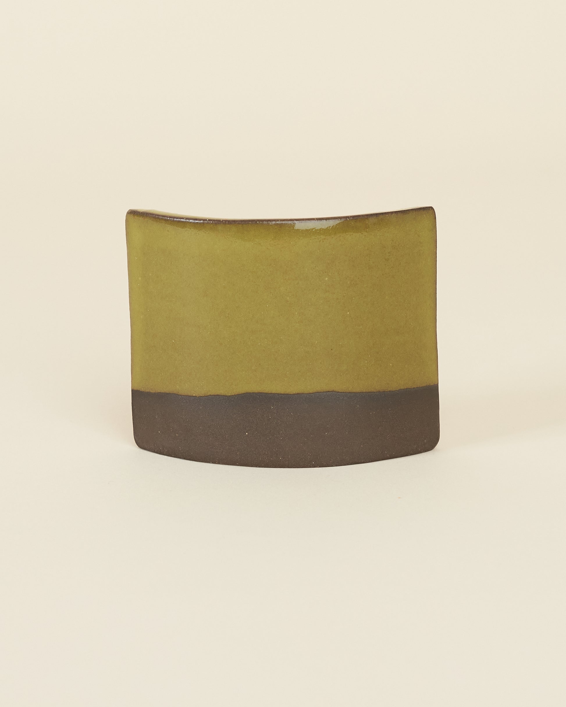 A curved square test tile made of dark brown clay and a glossy chartreuse glaze sits on a cream background.