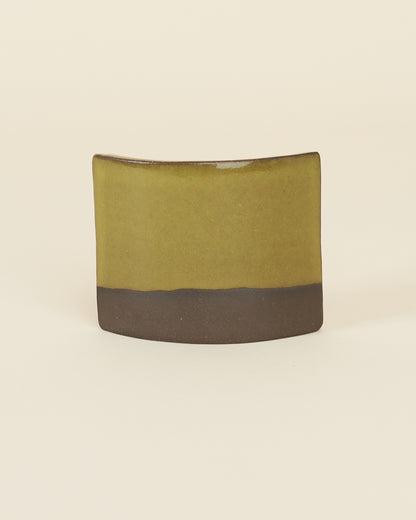 A curved square test tile made of dark brown clay and a glossy chartreuse glaze sits on a cream background.