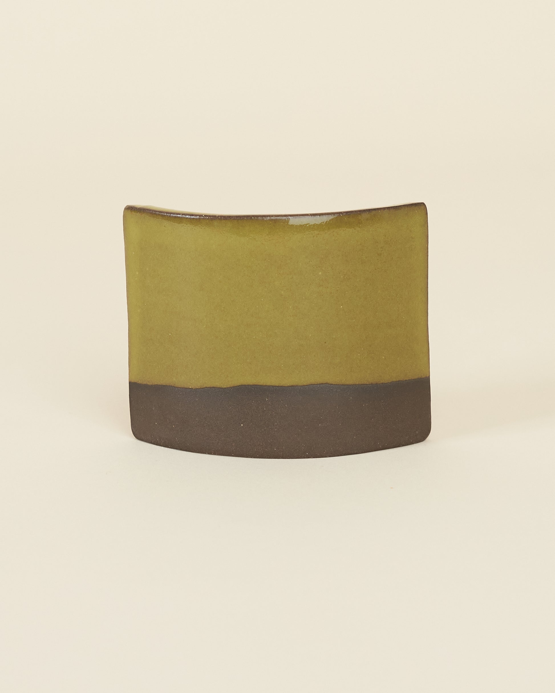 A curved square test tile made of dark brown clay and a glossy chartreuse glaze sits on a cream background.