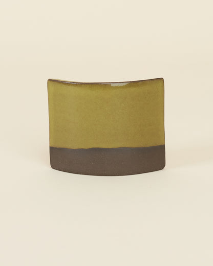A curved square test tile made of dark brown clay and a glossy chartreuse glaze sits on a cream background.