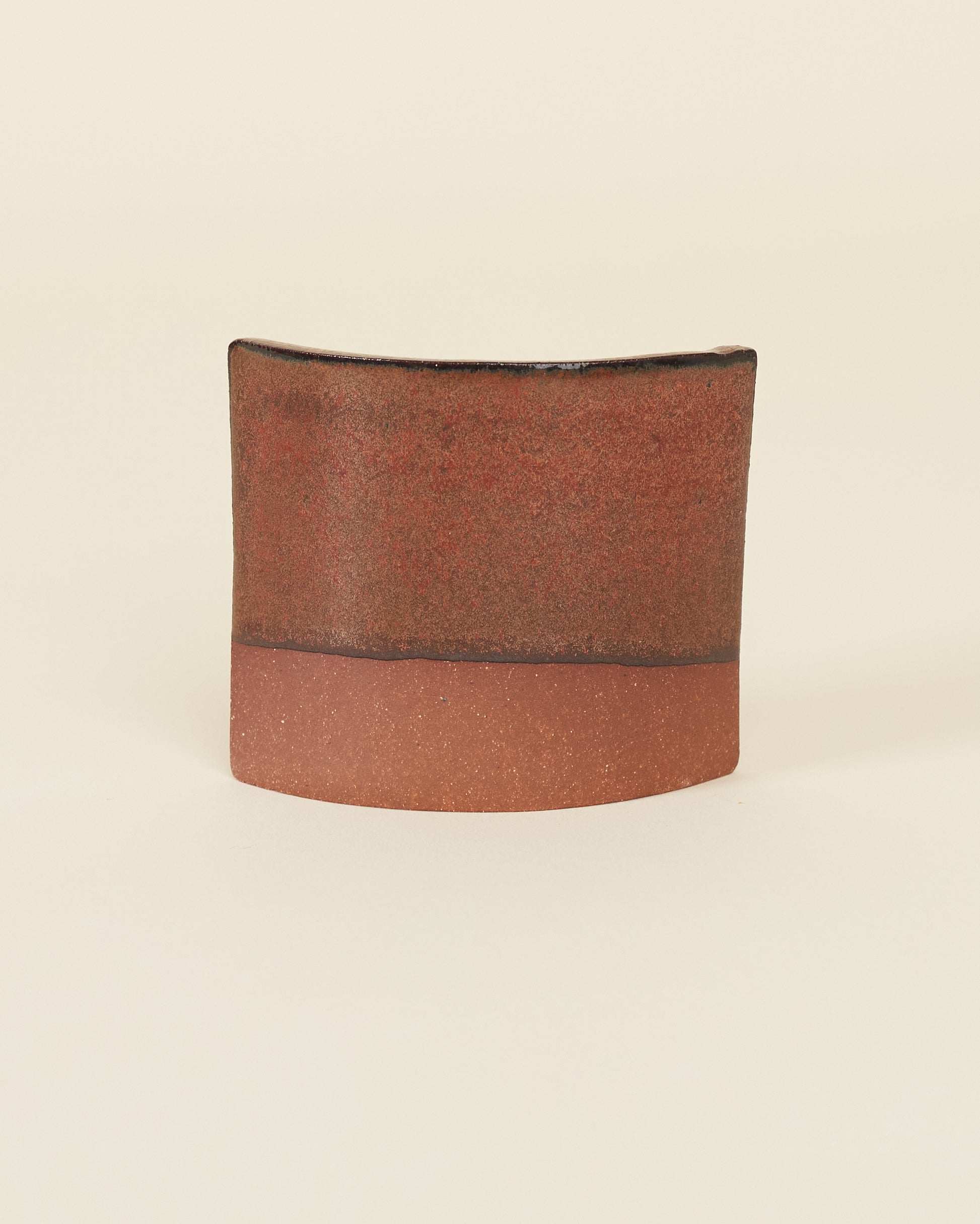 A curved square test tile made of red clay and a satin variegated red glaze that breaks black on the edges sits on a cream background.
