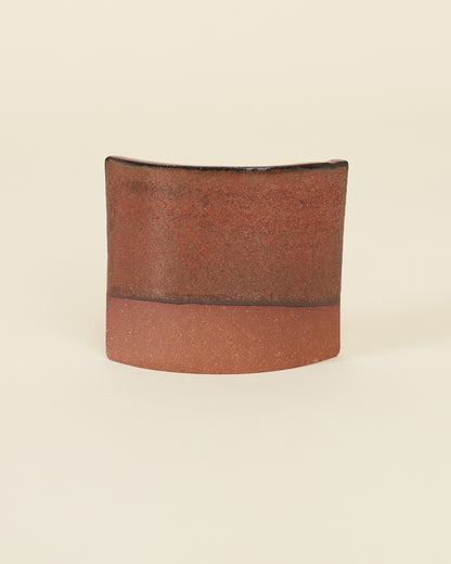 A curved square test tile made of red clay and a satin variegated red glaze that breaks black on the edges sits on a cream background.