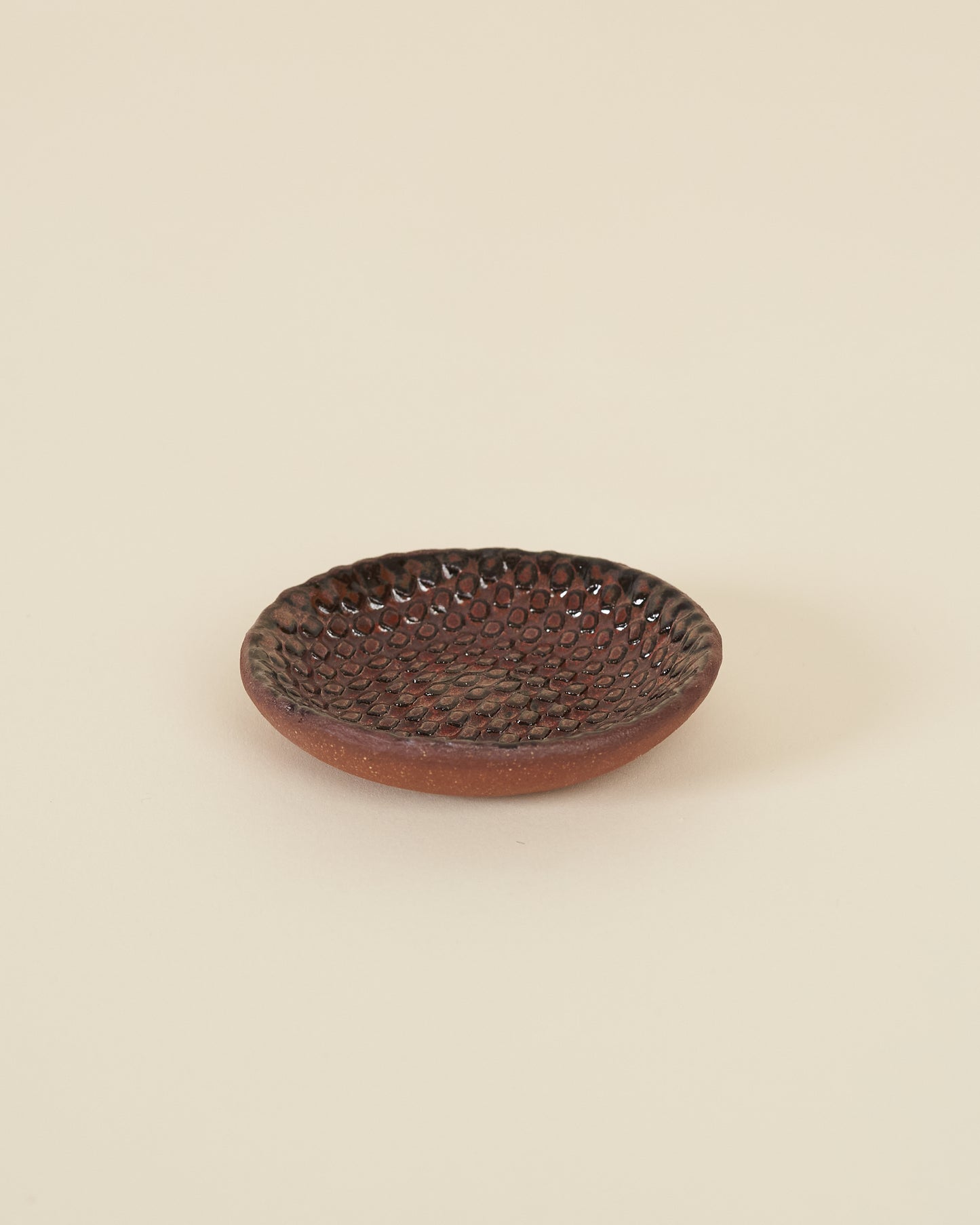 A small checkerboard textured dish made of red clay and a dark satin red glaze sits on a cream background.