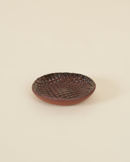 A small checkerboard textured dish made of red clay and a dark satin red glaze sits on a cream background.