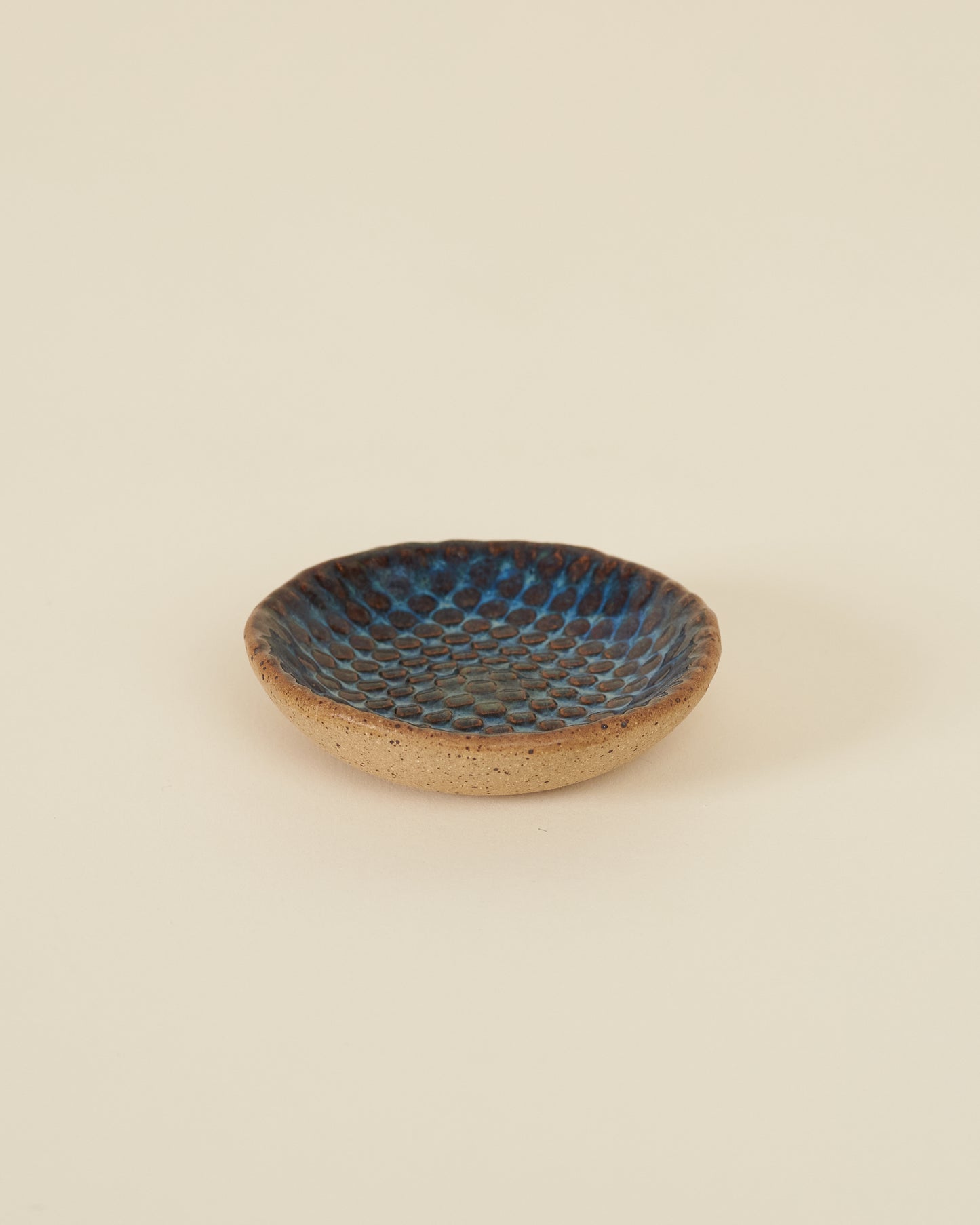 A small checkerboard textured dish made of speckled tan clay and a rich glossy blue variegated glaze sits on a cream background.