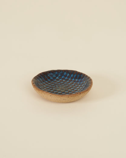 A small checkerboard textured dish made of speckled tan clay and a rich glossy blue variegated glaze sits on a cream background.