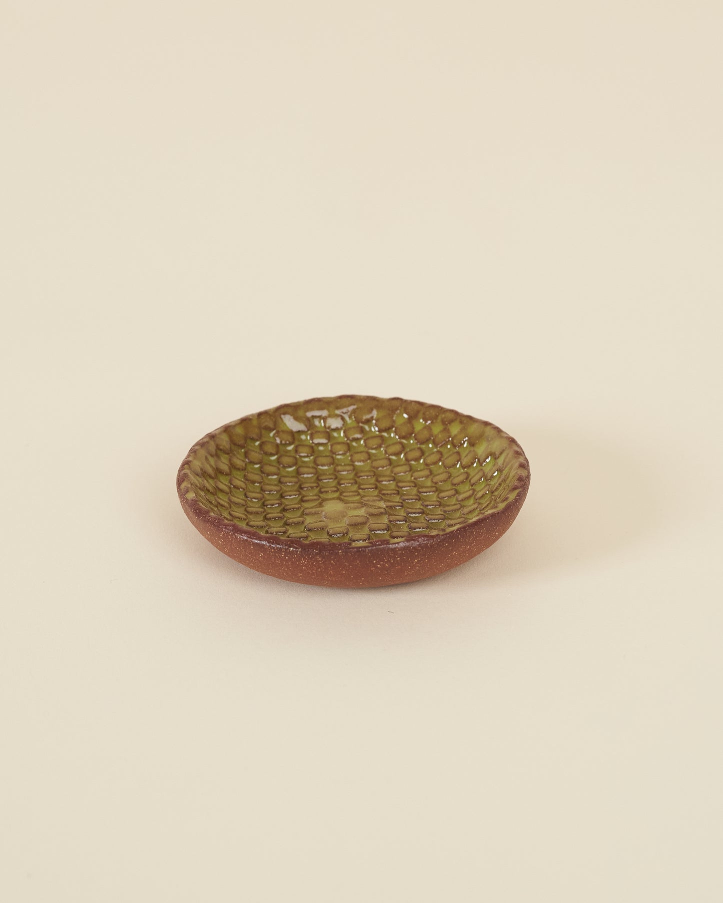 A small checkerboard textured dish made of red clay and a glossy chartreuse glaze sits on a cream background.