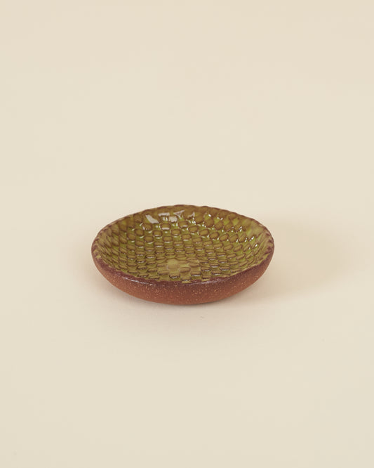 A small checkerboard textured dish made of red clay and a glossy chartreuse glaze sits on a cream background.